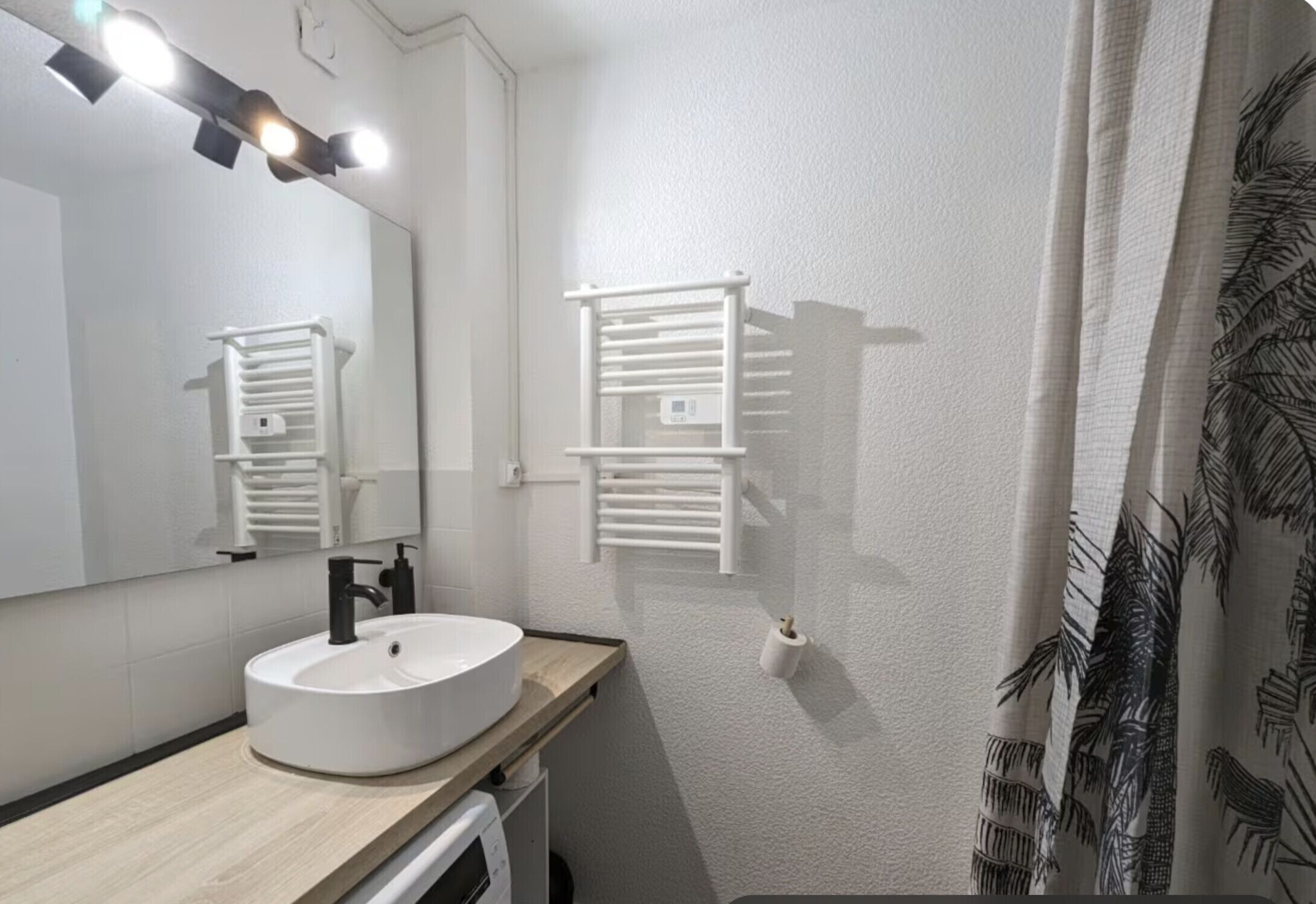 Une salle de bain avec un lavabo blanc, un miroir, un porte-serviettes et un rideau de douche orné d’un motif de feuilles.