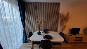 Un coin repas comprenant une table, des chaises, un vase avec des plantes séchées, un téléviseur posé sur un meuble et une fenêtre avec des rideaux.