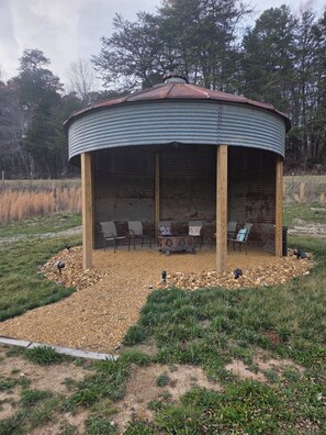 Un mirador con techo de metal corrugado, postes de madera y una zona de estar con sillas y una mesa.