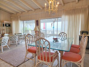 A dining area with a glass table and wicker chairs, a chandelier, and a view of the ocean. - Trident Ocean Front & Pet Friendly (Pawleys Island)