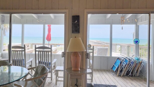 Ein Zimmer mit Glastür, die auf eine Terrasse mit Blick zum Strand führt, einem Tisch mit Stühlen und einer Lampe.