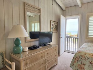 A room with a television on a dresser, a lamp, a framed picture, and a door leading to a balcony with a view. - Trident Ocean Front & Pet Friendly (Pawleys Island)