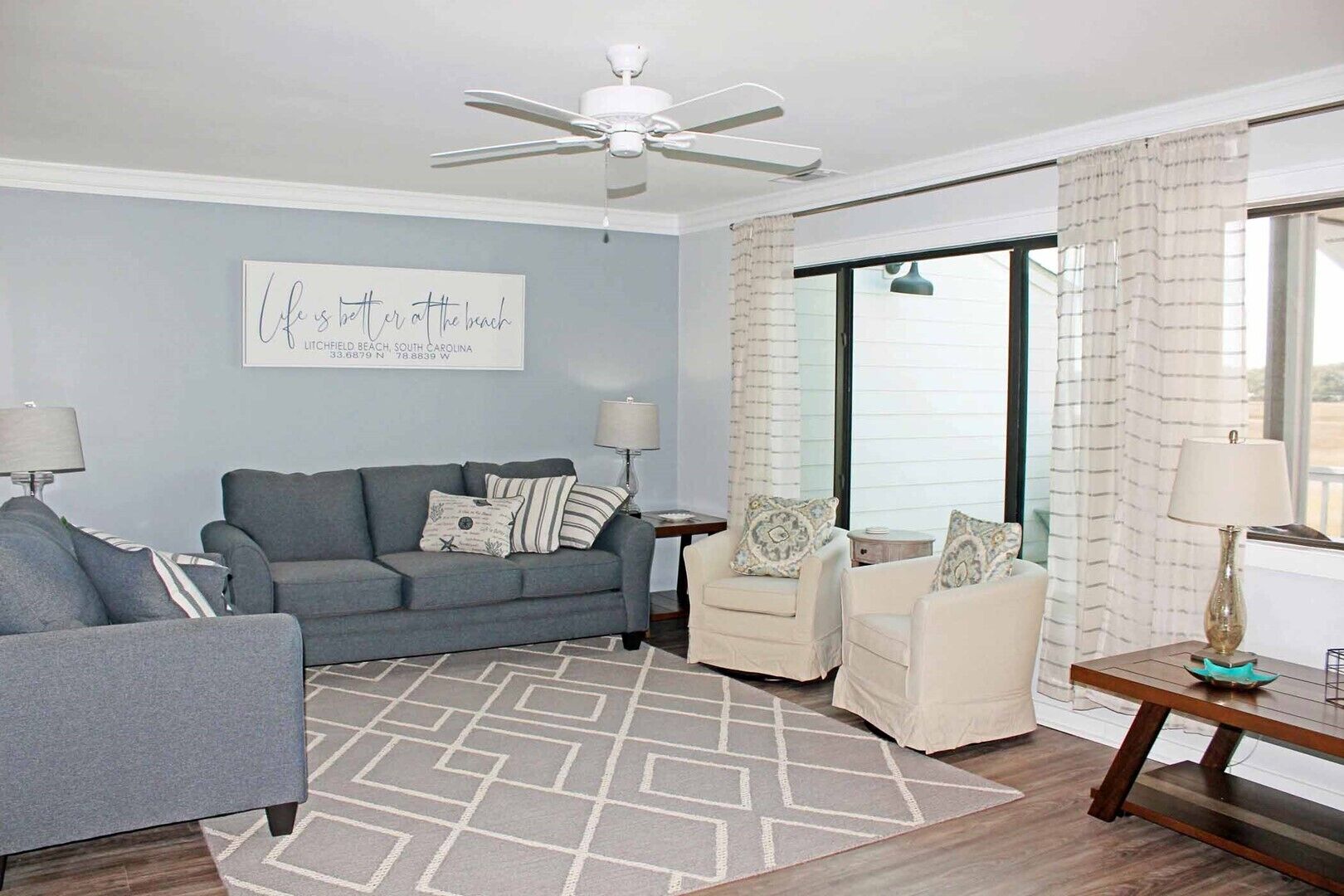 A living room with a grey sofa, a white armchair, a patterned rug, a ceiling fan, and a window with curtains.