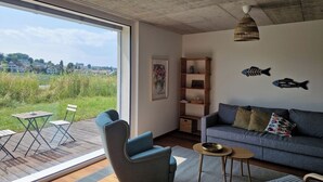 Ein modernes Wohnzimmer mit großem Fenster, einer Couch, einem Sessel, einem kleinen Tisch und einem Bücherregal.