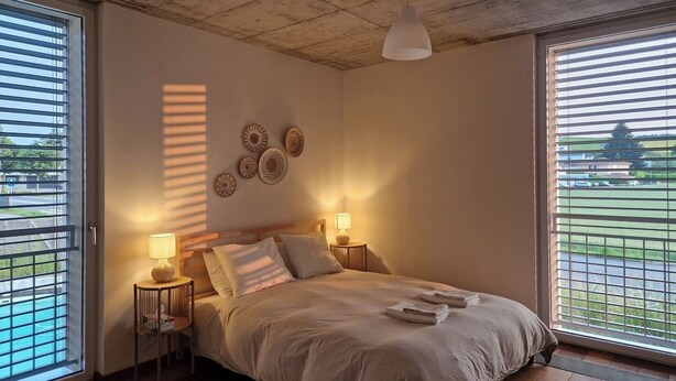 Ein Schlafzimmer mit einem Bett, zwei Nachttischlampen, einem Deckenventilator und Blick durch große Fenster.