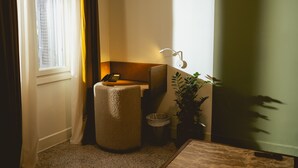 Una esquina de habitación con ventana, un taburete, un cubo de basura, una planta y una lámpara.