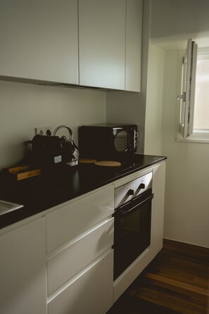 Una cocina con microondas, hervidor y tabla de cortar de madera sobre la encimera, armarios blancos y una ventana.