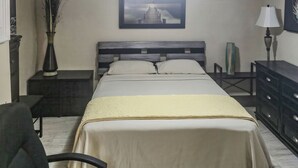 A bedroom with a bed, a ceiling fan, a nightstand with a lamp, a vase with plants, and a painting on the wall. - Studio Efficiency Apartment (Port Saint Lucie)
