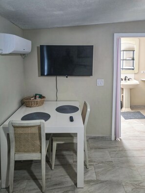 A dining area with a table, chairs, a television, and a wall-mounted air conditioner. - Studio Efficiency Apartment (Port Saint Lucie)