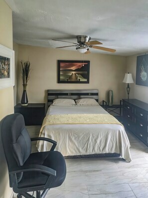 A bedroom with a bed, a desk, a chair, a ceiling fan, and artwork on the walls. - Studio Efficiency Apartment (Port Saint Lucie)