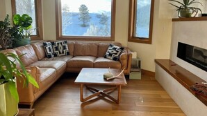 A living room with a brown leather sofa, a wooden coffee table, and a fireplace. There are three windows with views of a snowy landscape. - Downtown Oasis with View, Hot Tub & Fire Pits (Pagosa Springs)