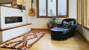 A living room with a fireplace, a large window with a view of trees, a guitar, and a black leather chair. - Downtown Oasis with View, Hot Tub & Fire Pits (Pagosa Springs)