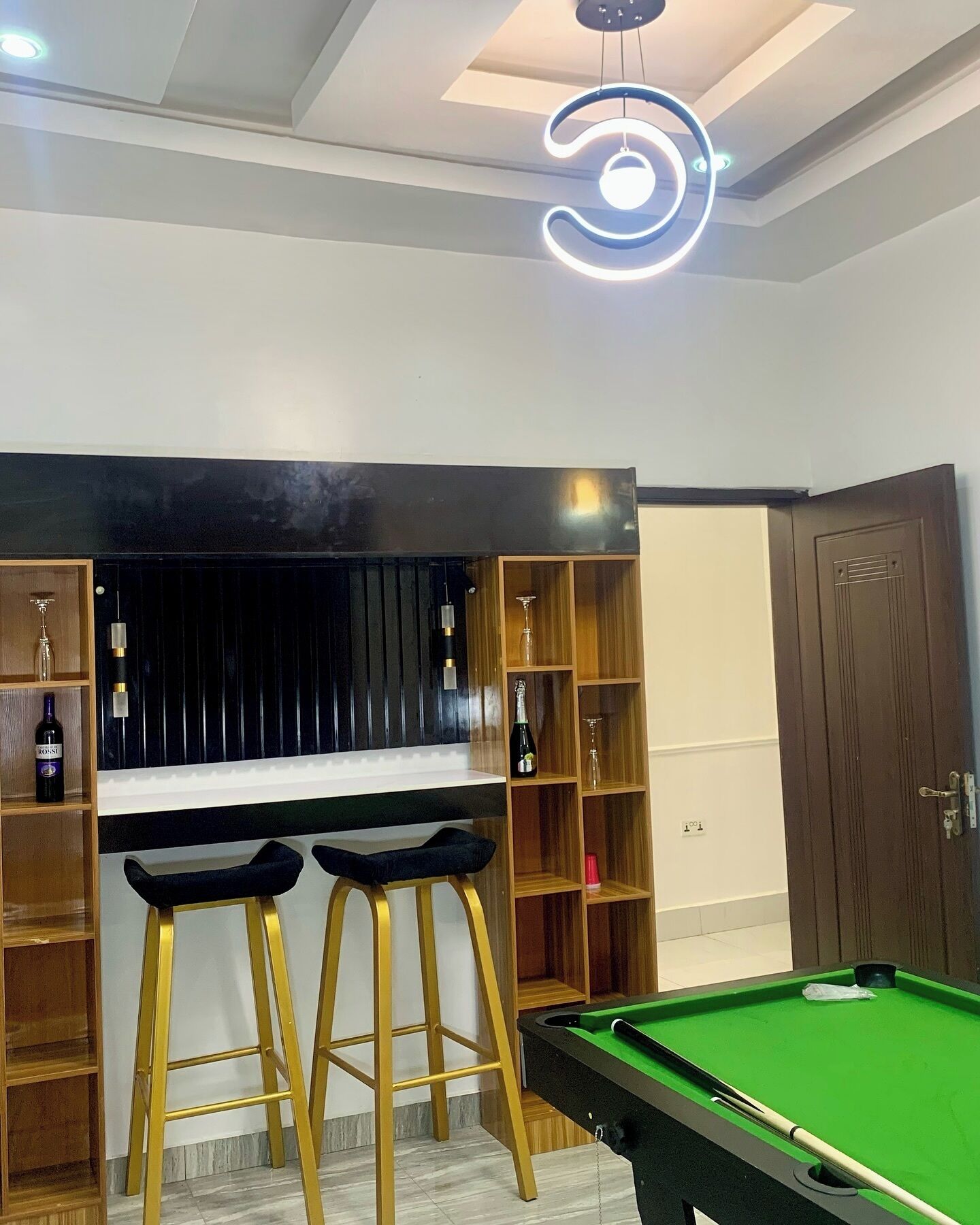 Une table de billard recouverte d’un drap vert, deux tabourets aux pieds dorés, un coin bar avec des étagères et un luminaire au plafond.