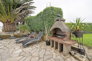 Une terrasse extérieure avec un sol en pierre, un barbecue en pierre et des chaises longues.