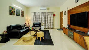 A living room with a black leather sofa, a coffee table, a TV, and a cabinet. There is a window with curtains, an air conditioner, and wall art. - Alaya Stays Saffron Breeze 4 BHK Apt (Bengaluru)