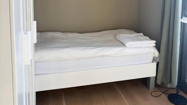 Een kleine eenpersoonskamer met een houten vloer, een wit bed en een gordijn voor het raam.