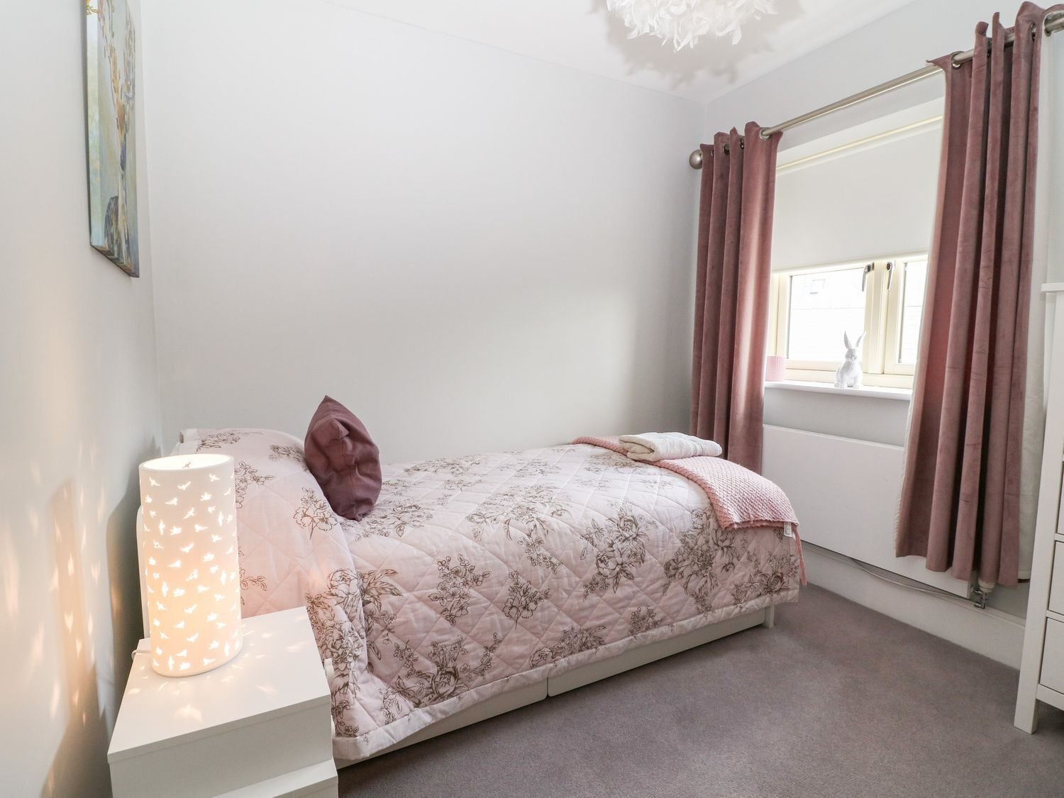 Un dormitorio con cama, lámpara de noche, ventana con cortinas e imagen colgada en la pared.