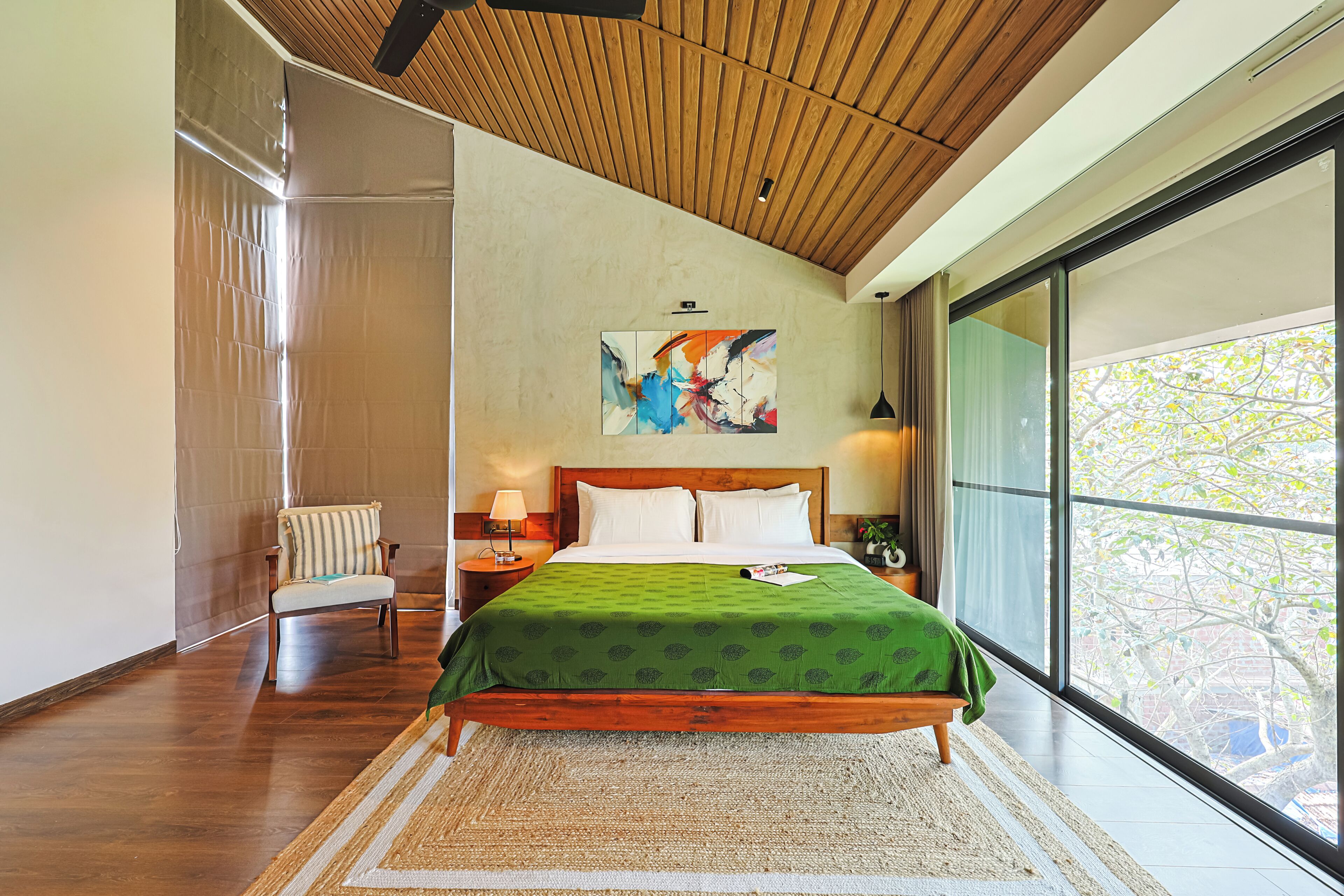A modern bedroom with a wooden ceiling, a large bed with a green bedspread, a chair, a nightstand with a lamp, and a painting on the wall.