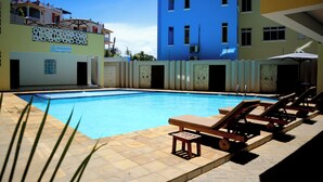 A swimming pool with lounge chairs and a small table, surrounded by buildings with balconies and a sign that reads 'ZUNI BAHAM'.