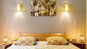 A bedroom with a wooden headboard, two bedside tables with lamps, a bed with patterned bedding, and a wall-mounted picture of a forest scene. - Gîte Douceur de Vivre - Marais: Lake View, Shared Pool & Wi-Fi (Saint-Avaugourd-des-Landes)