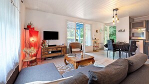 A modern living room with a sofa, a coffee table, a TV, and a dining area with chairs and a table. - Gîte Douceur de Vivre - Marais: Lake View, Shared Pool & Wi-Fi (Saint-Avaugourd-des-Landes)