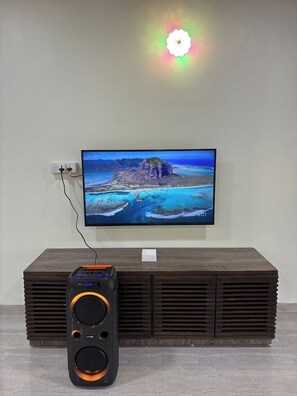 A flat-screen TV mounted on a wall, a speaker in front of it, and a power outlet to the left. - Scenic Karjat Retreat – 4BHK Riverstone Villa with Pool & Nature Surroundings (Karjat)