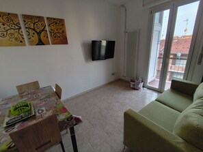 A living room with a sofa, a table with chairs, a TV mounted on the wall, and a sliding glass door leading to an outdoor balcony.