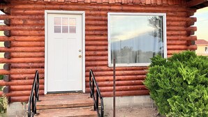 A log cabin with a white door and window, wooden steps, and a metal railing.