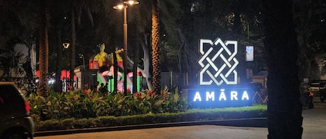 A nighttime view of a sign with the word 'AMARA' illuminated, surrounded by greenery and palm trees.