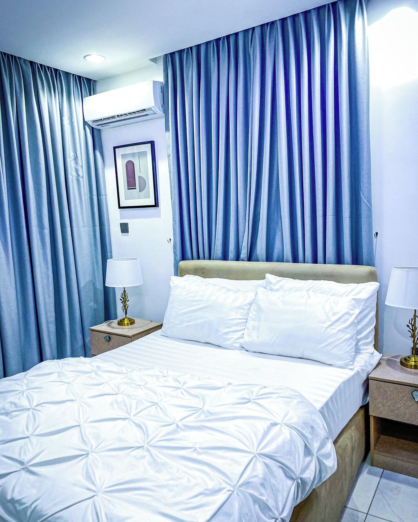 A hotel room with a bed, curtains, a wall-mounted air conditioner, a framed picture, and bedside lamps.