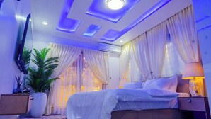 A bedroom with a bed, curtains, a TV, a plant, and a nightstand. - THE 81 APARTMENTS (Lagos)
