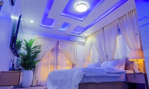 A bedroom with a bed, curtains, a TV, a plant, and a nightstand. - THE 81 APARTMENTS (Lagos)