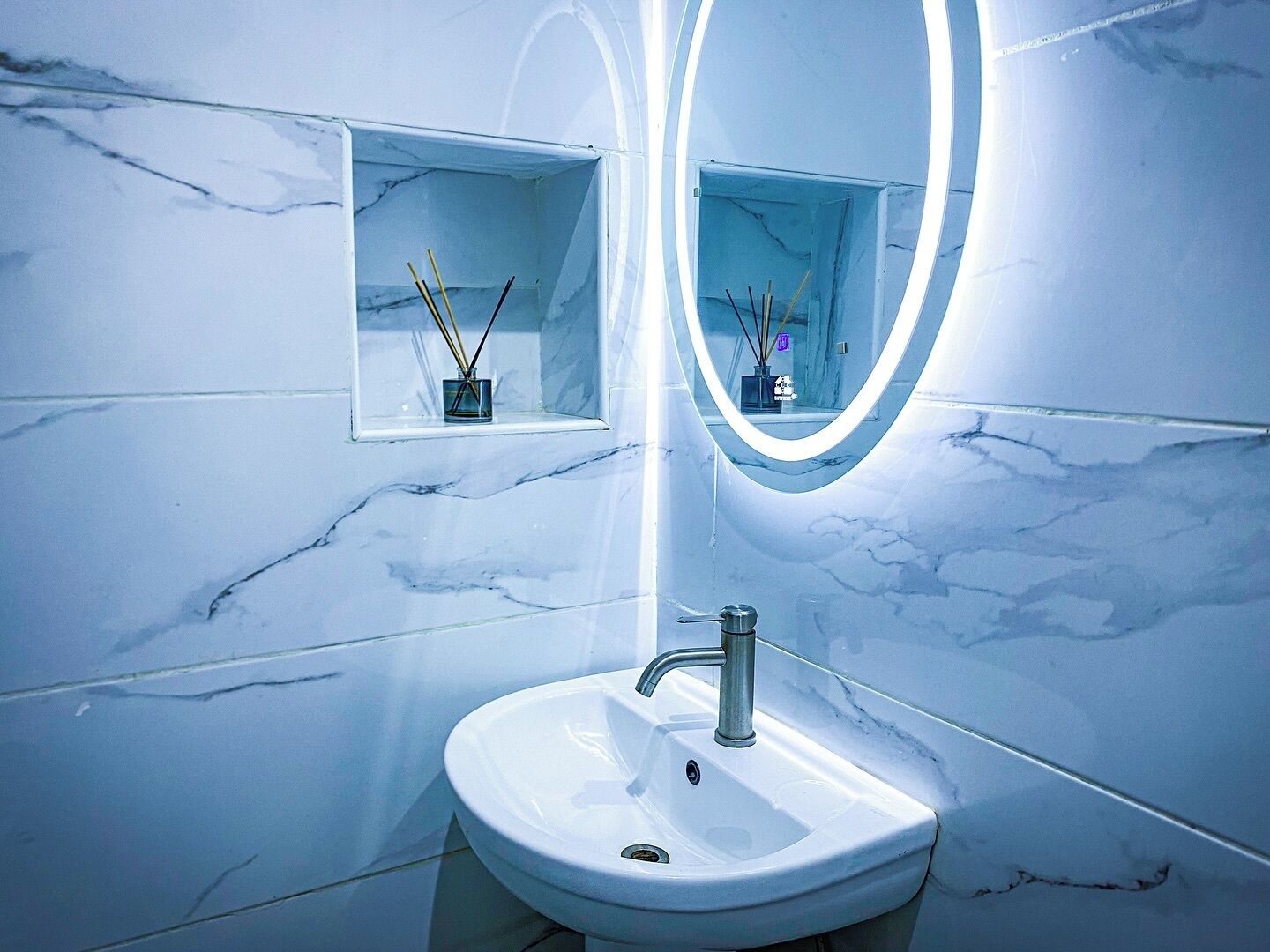 A bathroom with a white sink, a mirror with a light, and two small shelves with reed diffusers.