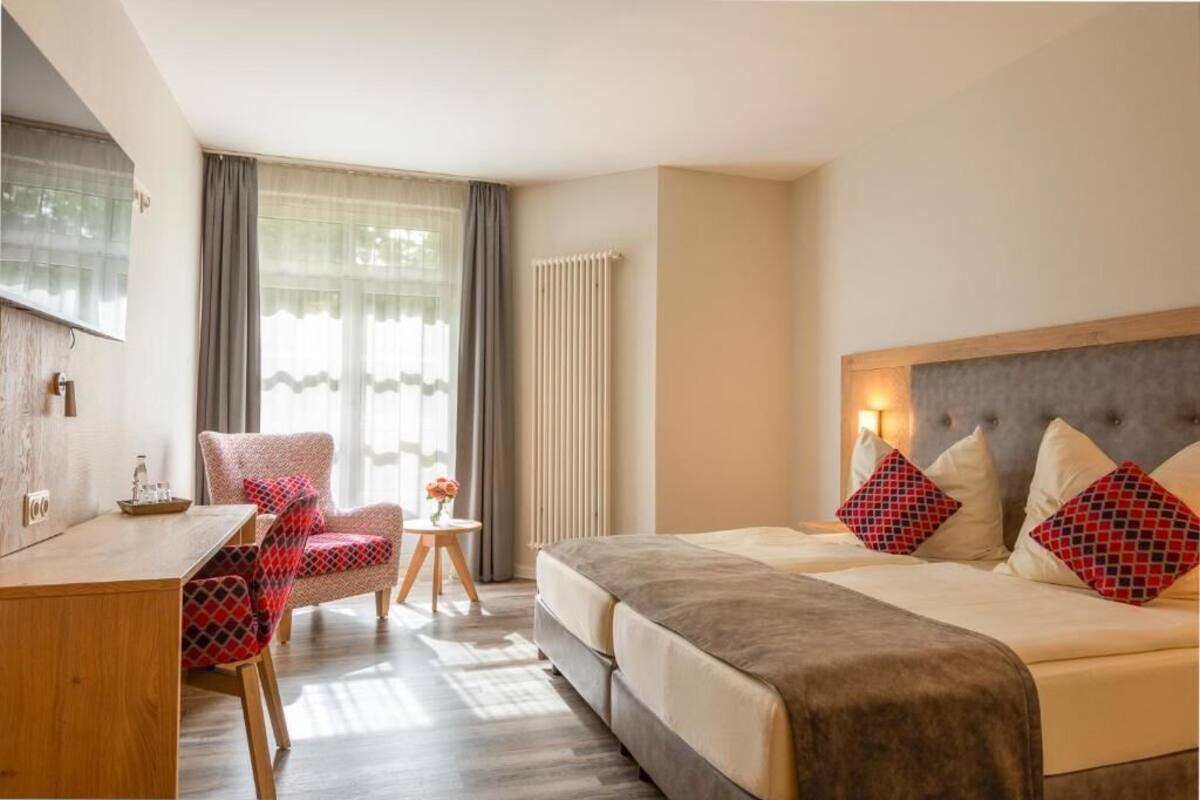 A hotel room with a bed, a desk, a chair, a small table with a vase, and a radiator.