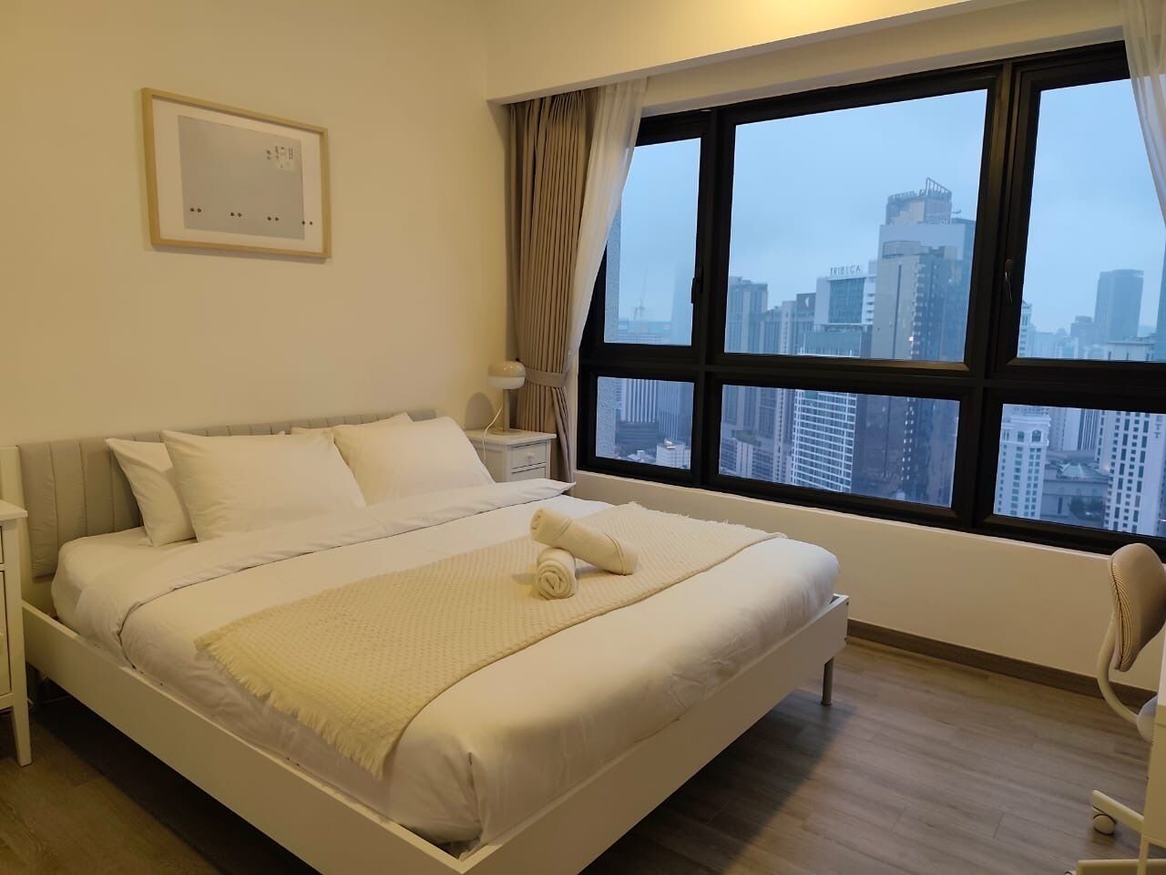 A hotel room with a large bed, a nightstand, a chair, and a view of city buildings through the window.