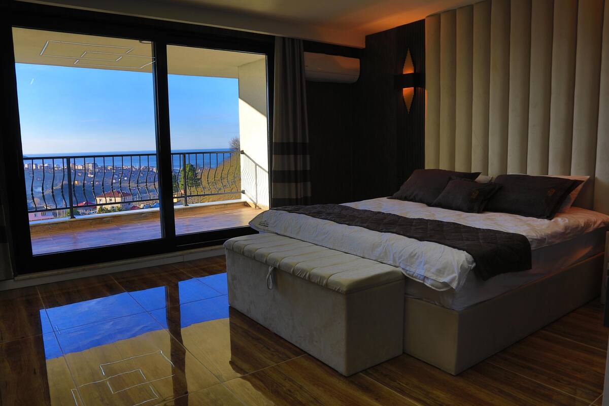 A bedroom with a large bed, a bench, and a balcony with a view.