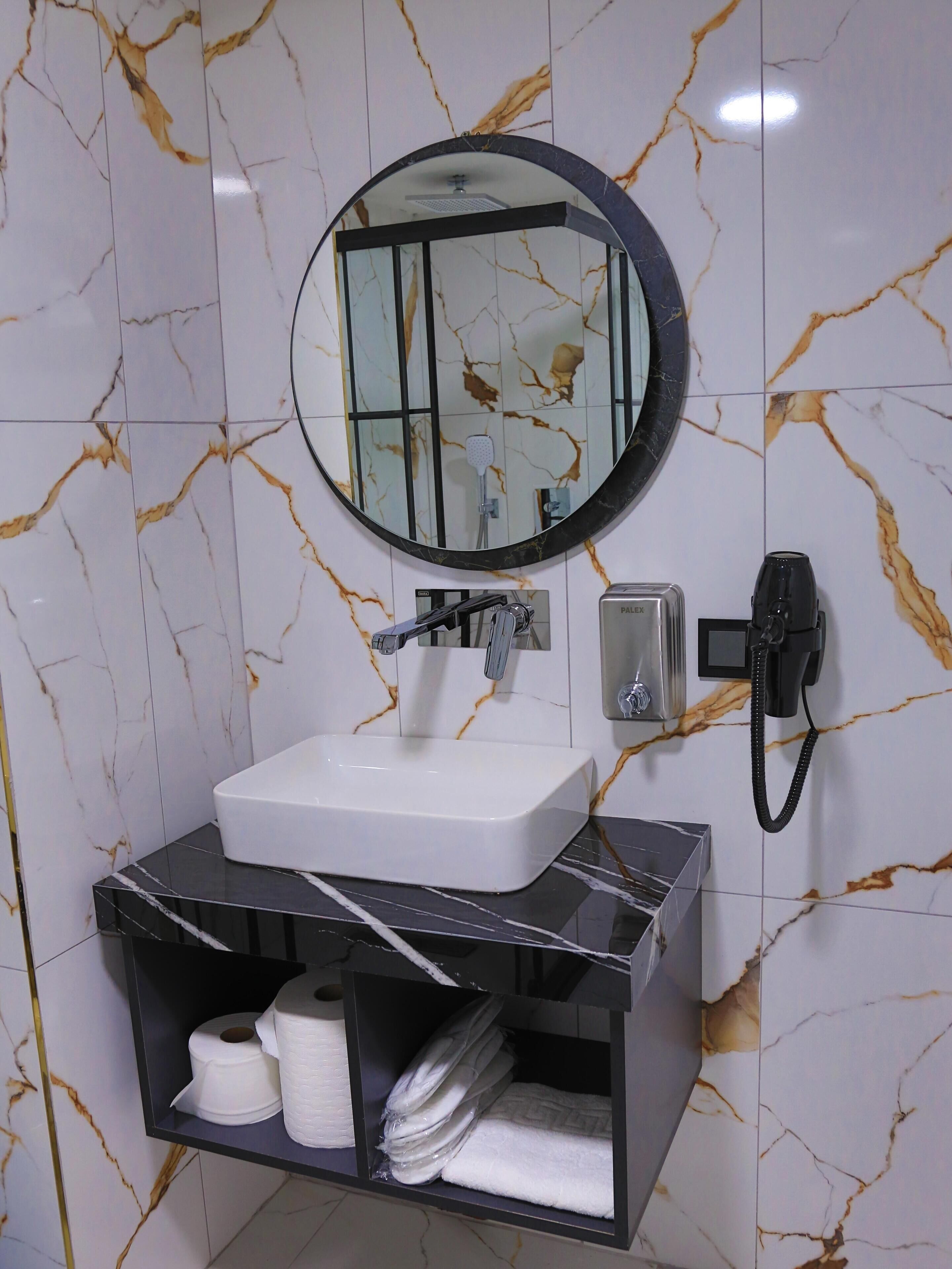 A bathroom with a white sink, a round mirror, a handheld showerhead, a toilet paper holder, and a towel rack.