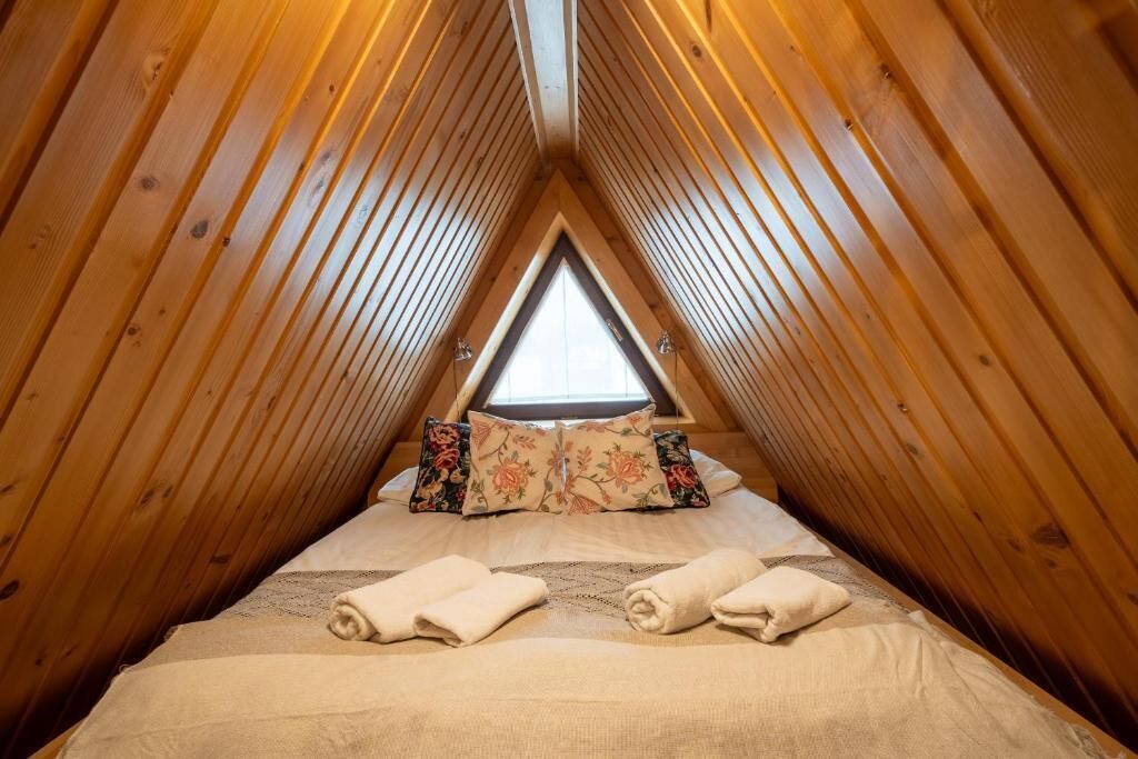 Une chambre confortable située dans les combles, avec des murs en bois, une fenêtre triangulaire et un lit agrémenté d’oreillers à motifs floraux.