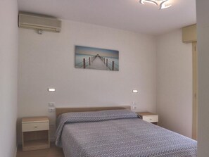 A bedroom with a bed, a nightstand, a wall-mounted air conditioner, and a painting of a pier. - Cozy Apartment With two Terraces (Lignano Sabbiadoro)