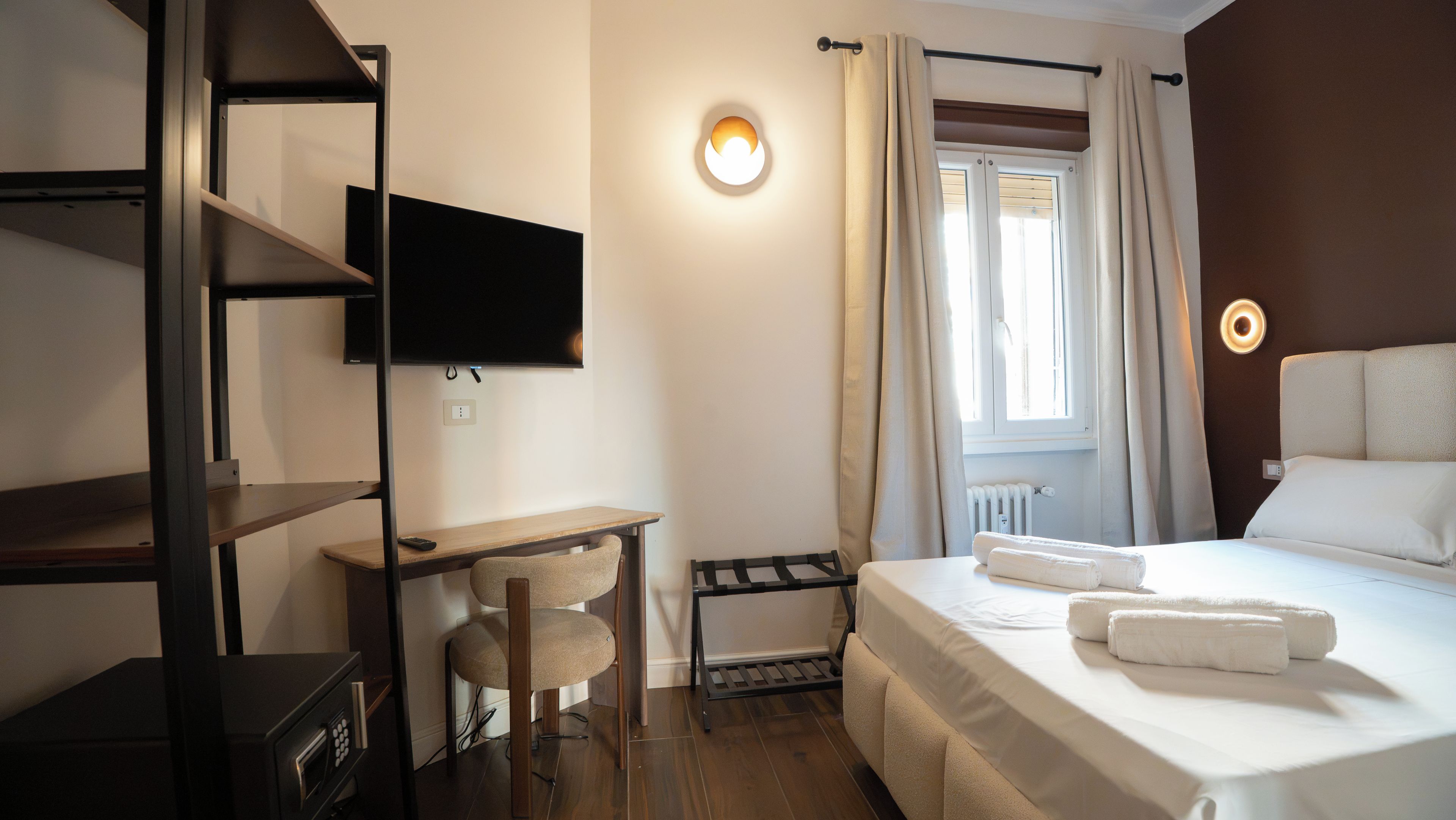 A hotel room with a bed, a desk, a chair, a TV mounted on the wall, and a window with curtains.