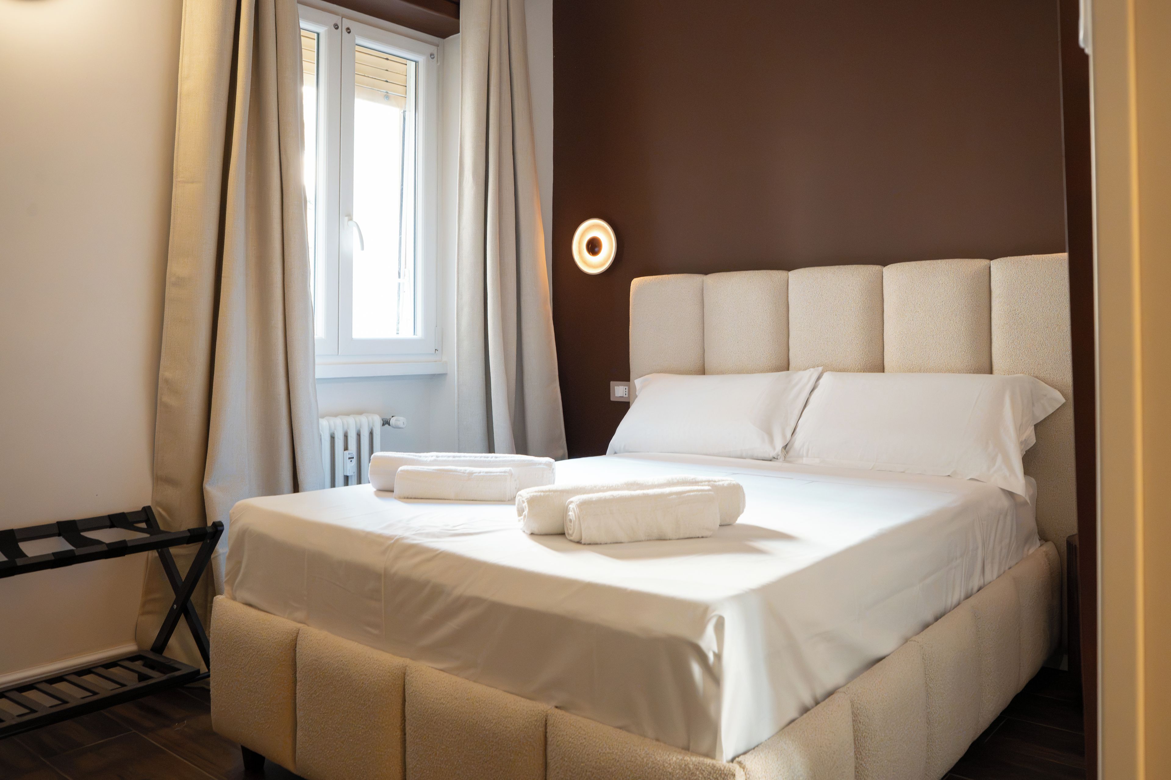 A neatly made bed with white linens and two rolled towels, a bedside table with a lamp, and a window with curtains.