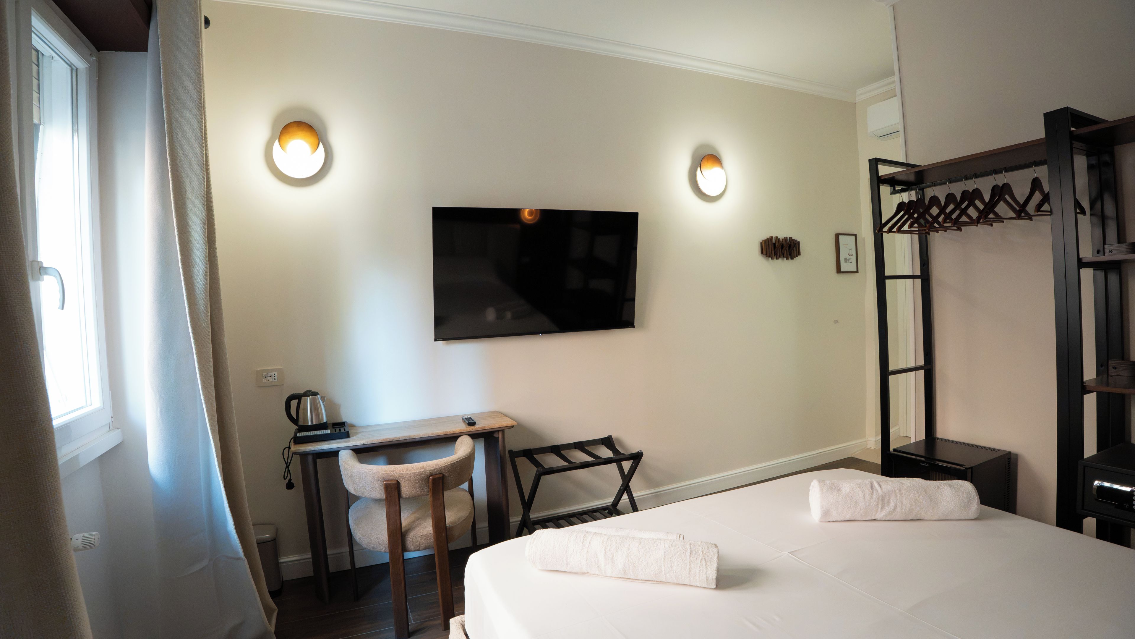 A hotel room with a bed, a desk with a chair, a TV mounted on the wall, and a coat rack.