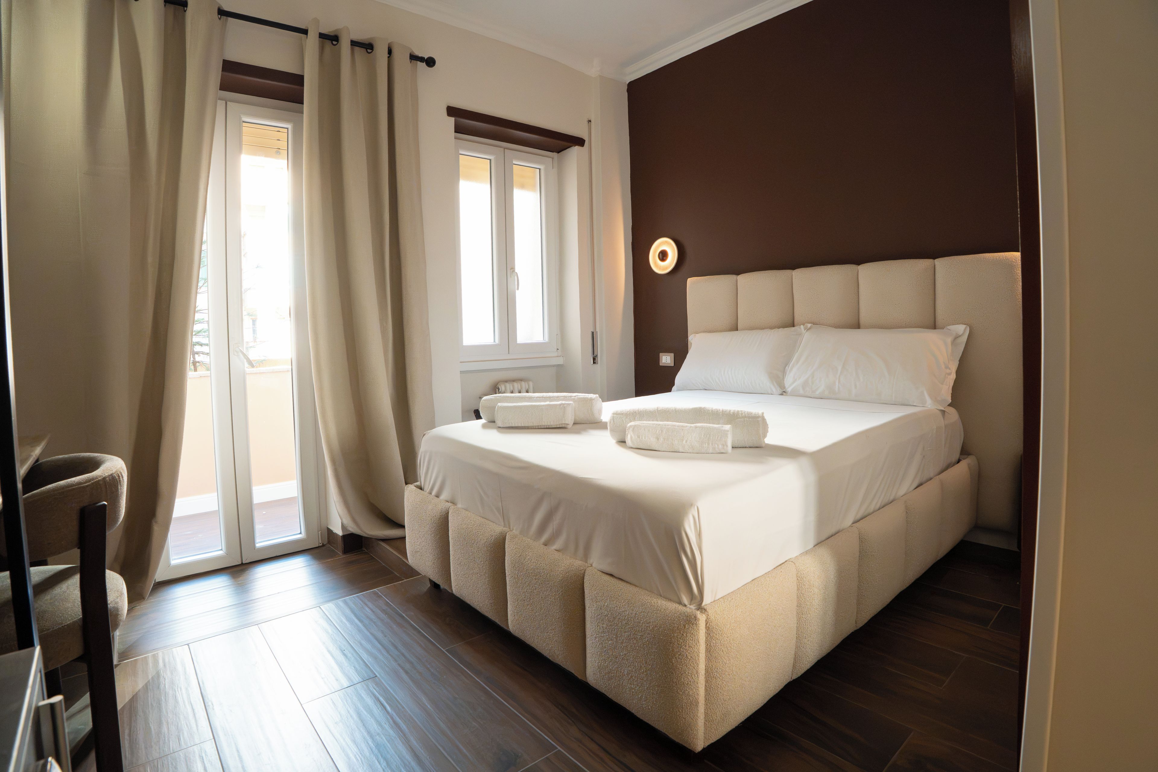 A hotel room with a bed, a chair, a window with curtains, and a wall-mounted light.