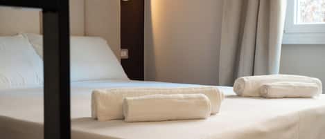 Una cama bien hecha con sábanas blancas y dos toallas enrolladas, una lámpara de noche y una ventana con cortinas.