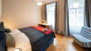 A modern hotel room with a large bed, a desk, a chair, and a view of the city through the windows. - DOWNTOWN - 2 Room Large Apartment (Praha)