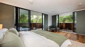 Ein modernes Schlafzimmer mit einem großen Bett, einer Sitzecke mit einem Sessel und einem kleinen Tisch sowie großen Fenstern mit Blick auf Grünflächen.