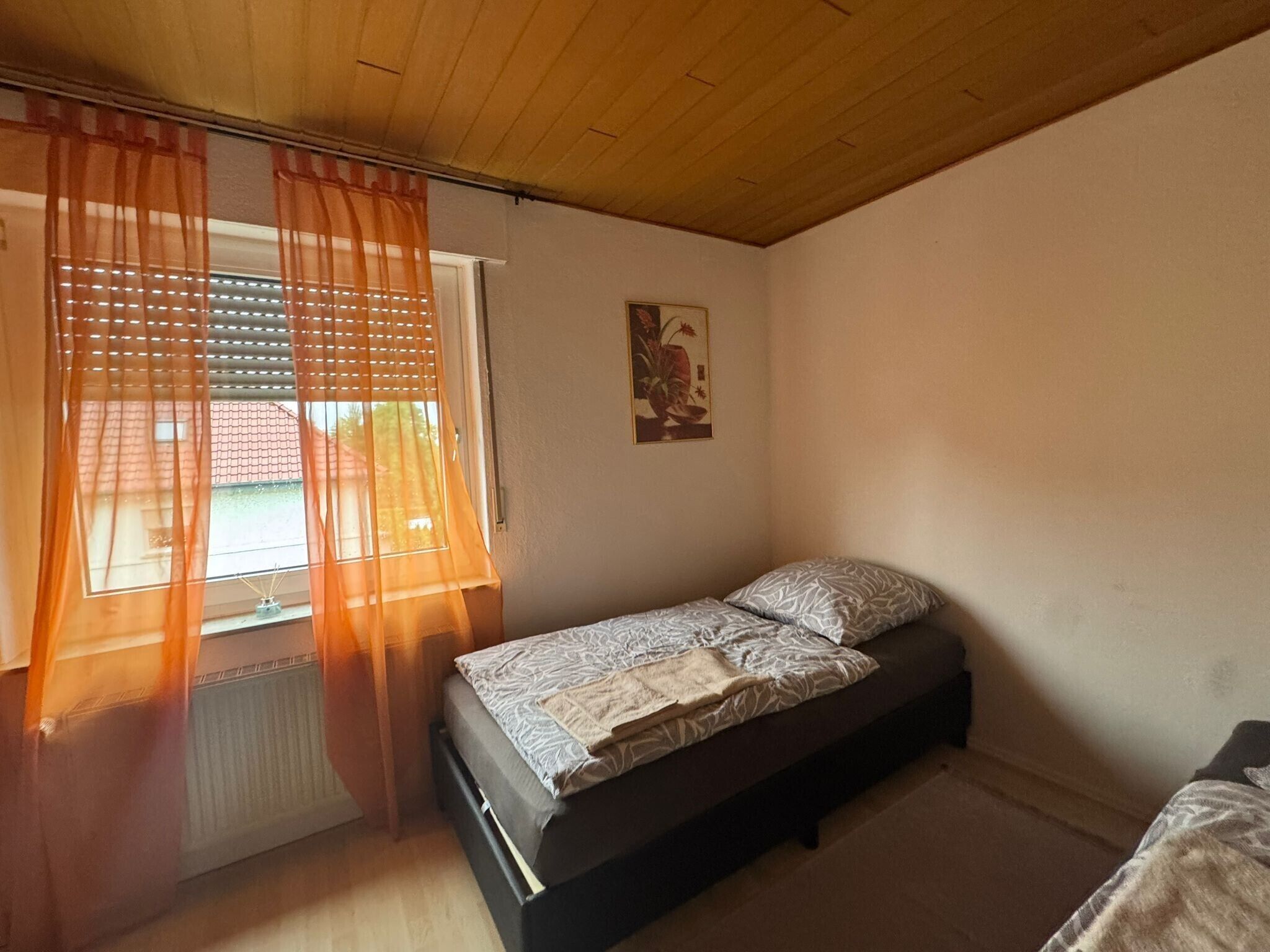 A small bedroom with a single bed, a window with orange curtains, a radiator, and a wall-mounted picture.