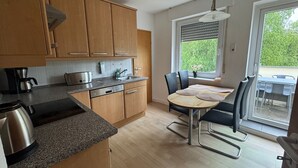 A modern kitchen with wooden cabinets, a dining table with chairs, a coffee maker, and a window with blinds. - Apartment 'Beautiful 100m² Holiday' with Private Terrace, Shared Garden and Wi-Fi (Herford)