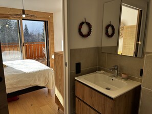 A bedroom with a bed, a wooden wardrobe, a bathroom with a sink and mirror, and a view of a deck with a wooden fence. - Modern apartment with parking - 8 guests (Arâches-la-Frasse)