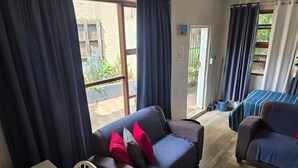 Hyggelig opholdsstue med sofa, lænestol og seng med stribet dyne. En skydedør fører til et udendørs område med planter.